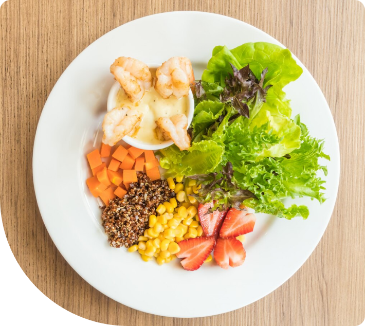 En fargerik salat med grillete reker i en hvit saus, sammen med quinoa, gulrøtter, mais, jordbær og ulike grønne salater, servert på en hvit tallerken.