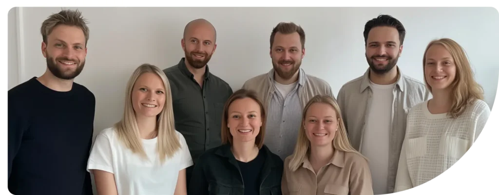 En gruppe på åtte personer, bestående av fem menn og tre kvinner, smiler mot kameraet. De står tett sammen foran en hvit vegg, kledd i en blanding av lyse og mørke klær.